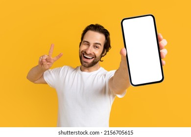 Cheerful emotional young european guy in white t-shirt show phone with empty screen and peace sign with hand isolated on yellow background, studio. Gesture, big sale, new app, device, app and offer