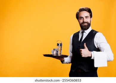 Cheerful employee with food tray gives like gesture in front of studio camera, showcasing approval or agreement sign. Positive butler shows thumbs up wearing formal attire in restaurant industry. - Powered by Shutterstock