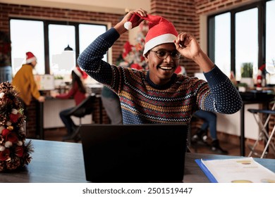 Cheerful employee putting santa hat on while working at desktop in festive decorated office at christmas eve and looking at camera. African american man at workplace during winter holiday season - Powered by Shutterstock