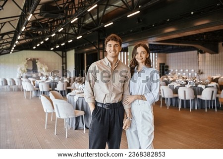 cheerful enamored couple smiling at camera near festive table in wedding venue, romantic setting