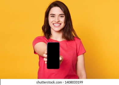 Cheerful energetic black haired female reaching out arm, holding smartphone in one hand, smiling sincerely, looking directly at camera, being in high spirits, wearing casual bright red t shirt. - Powered by Shutterstock