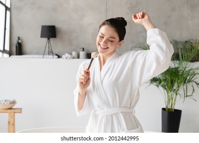 Cheerful energetic young woman in spa bathrobe singing and dancing using cosmetics brush as a microphone in the morning at home. Makeup and beauty treatment