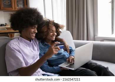 Cheerful Engaged Teenage Black Couple Shopping Online From Home, Using Laptop Computer, Plastic Credit Card, Resting, Hugging On Couch. Satisfied Bank Clients Making Payment, Buying On Internet