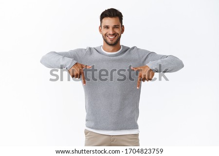Cheerful, enthusiastic active hispanic bearded man in grey sweater, pointing fingers down and smiling, give advice where find good product, promote application or store banner, white background