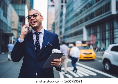 Cheerful Entrepreneur In Trendy Suit And Sunglasses Answering Phone Call Next To Intersection Holding Tablet And Beverage In Hands On City Street 