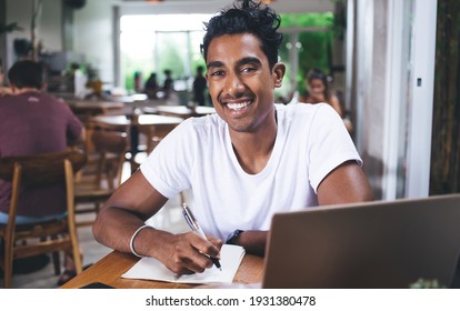 Cheerful ethnic man in casual t-shirt looking at camera while writing in planner and sitting at table with laptop in exotic cafeteria