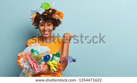 Cheerful ethnic woman with curly hair, carries trash, points away with thumb, picks up litter, reduces plastic wastes cares of envirnoment stands over blue background involved in campaing for cleaning