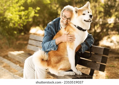 Cheerful european senior man in casual enjoys walk with dog, hugs, sits on bench in park, outdoor. Pet love, best friend care and free time in city, active lifestyle with animal - Shutterstock ID 2310519037