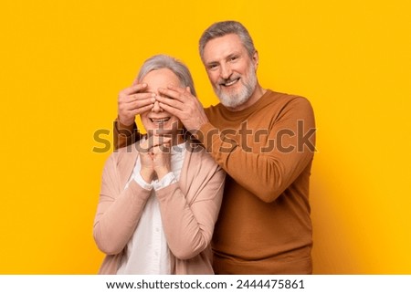 Cheerful European senior man closes eyes to his wife smiling to camera, preparing anniversary or birthday surprise, standing against yellow studio background. Romantic Surprises, Celebration