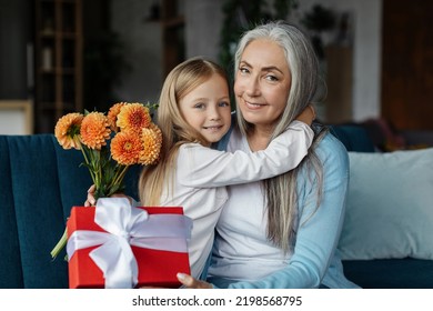 Cheerful European Small Girl Hugging Old Woman With Gift Box And Bouquet Of Flowers In Living Room Interior. Grandma And Granddaughter Relationship, Congratulations On Holiday And Birthday Celebration