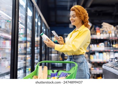 Cheerful European woman scanning qr code of daily product using smartphone, standing near refrigerator. Concept of shopping and consumerism.