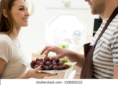 Cheerful excited beautiful woman in t-shirt giving fruits to husband and looking at him, smiling man taking grape from bowl at home