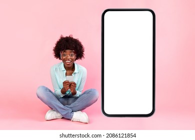 Cheerful excited black lady sitting on floor near big cellphone with empty white screen, using mobile device, checking new cool app on pink studio background, mock up for website or application design