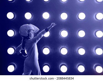 Cheerful Excited Child In White Spacesuit And Helmet Standing With Arm Raised Against Spaceship Wall With Glowing Round Glowing Lamps. Ttoned