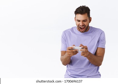 Cheerful, Excited Good-looking Hispanic Bearded Guy In Purple T-shirt, Looking Astonished And Thrilled, Playing Awesome New Smartphone Game, Yelling And Smiling Pleased, Winning, White Background