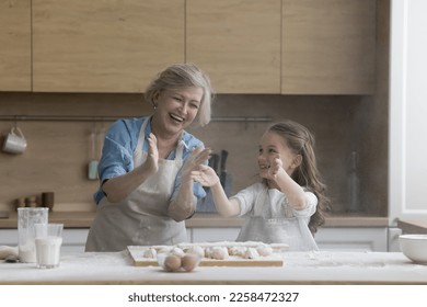 Cheerful excited grandmother and granddaughter kid having fun in home kitchen, baking, hitting floury hands over table with dough, making cloud of flour, laughing