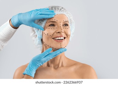 Cheerful excited middle aged woman in medical hat with pre surgery marks on her face looking at copy space for ad and smiling, surgeon hands in blue gloves touching female skin