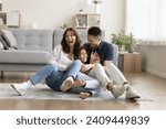 Cheerful excited parents tickling beloved kid girl, playing with child on heating floor in modern home interior, sitting on carpet, laughing, shouting for joy, enjoying family leisure