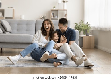Cheerful excited parents tickling beloved kid girl, playing with child on heating floor in modern home interior, sitting on carpet, laughing, shouting for joy, enjoying family leisure