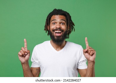 Cheerful excited young african american man guy with dreadlocks 20s in white casual t-shirt posing pointing index fingers up on mock up copy space isolated on green color background studio portrait - Shutterstock ID 2142284027
