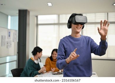 A cheerful and excited young Asian male developer tested a new VR game on a VR headset in the meeting with his team. Virtual reality, tech company, business startup