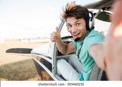 Cheerful excited young pilot sitting in cabin of small airplane and showing thumbs up