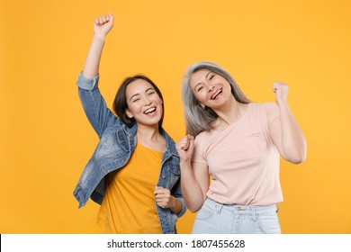 Cheerful Family Asian Women Gray-haired Mother, Daughter In Casual Clothes Posing Dancing Clenching Fists Rising Hands Up Having Fun Doing Winner Isolated On Yellow Color Background Studio Portrait.