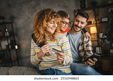 A cheerful family enjoy online shopping, parents and son smile as they share a moment of togetherness while holding a credit card in a cozy setting - Powered by Shutterstock