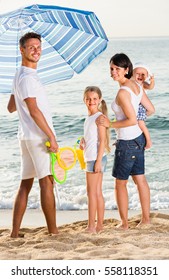 Cheerful Family Of Four Standing With Plastic Bucket And Scoops Under Sun Umbrella At Beach