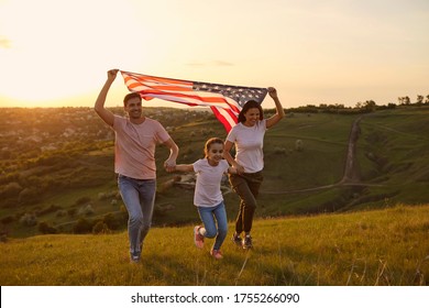 Cheerful Family Is Having Fun Running Along The Grass In A Summer Park. USA July 4th Celebration.