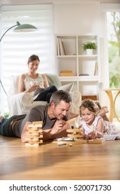 Cheerful Family At Home, Daddy And His Young Daughter Lying On The Wooden Floor Are Playing With A Wooden Game.  At Background Mom Is Sitting In An Armchair While Using A Digital Tablet