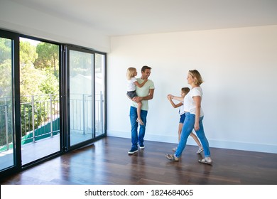 Cheerful family with kids looking around new home or apartment. Cute daughter sitting on father hands. Blonde mother in jeans pacing and going to balcony with girl. Relocation and moving day concept