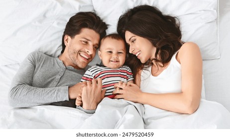 Cheerful family, a man and a woman, are lying in bed together, smiling at their baby. The baby is lying in the middle of the bed, between its parents, and is smiling back at them. - Powered by Shutterstock