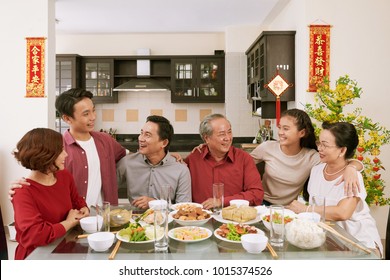 Cheerful Family Members Talking At Chinese New Year Celebration Dinner. Scrolls Wishing Luck And Wealth In The Background