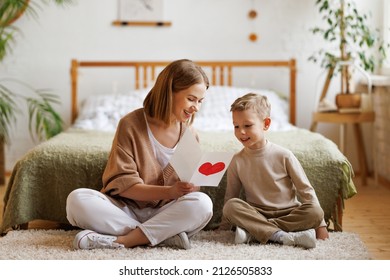 Cheerful family mom and  son and reading handmade greeting card with heart while resting on floor by bed during holiday celebration mothers day at home
