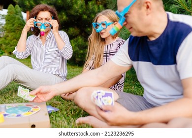 Cheerful Family Mother, Father And Their Daughter Playing Board Game While Sitting On Lawn In Backyard At Summer. Games And Entertainment For Children Concept.