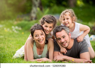 Cheerful Family In A Park, Dad, Mum And Heir Two Lovely Daughter Are Lying On The Grass While They Are Looking At Camera. Shot With Flare