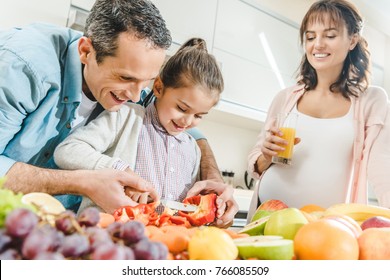 Cheerful Family, Pregnant Mother With Father Helping Daughter Slicing Pepper At Kitchen