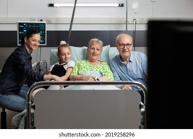 Cheerful family spending time with sick grandmother watching movie on tv during medical recovery visiting her in hospital ward. Senior woman patient resting in bed waiting for healthcare treatment