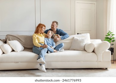 Cheerful family of three sitting on couch in light living room, parent and their daughter enjoy time together at home. Parenthood happiness - Powered by Shutterstock