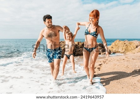 Cheerful family walking along seashore near waving ocean and enjoying vacation playing with kid on sunny day