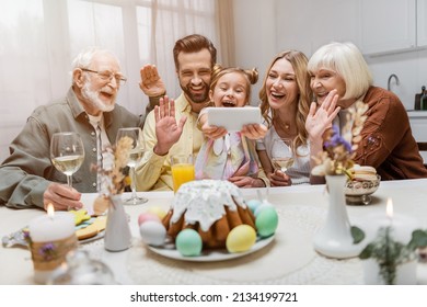 Cheerful Family Waving Hands During Video Call On Smartphone Near Table With Easter Dinner