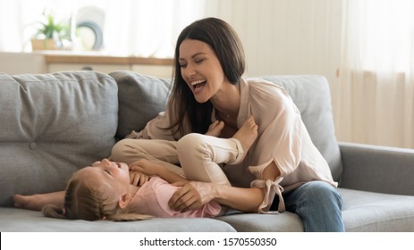 Cheerful family young adult mother tickling cute small child daughter laughing playing together on sofa in house living room, happy mom and little preschool kid having fun cuddle enjoy lifestyle game