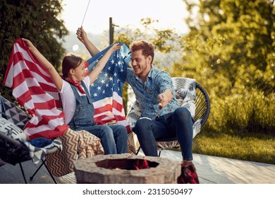 Cheerful family of young girl and father sitting outdoors in their yard roasting marshmallows and celebrating Independence day together, holding USA flag. Togetherness, holiday, family concept. - Powered by Shutterstock