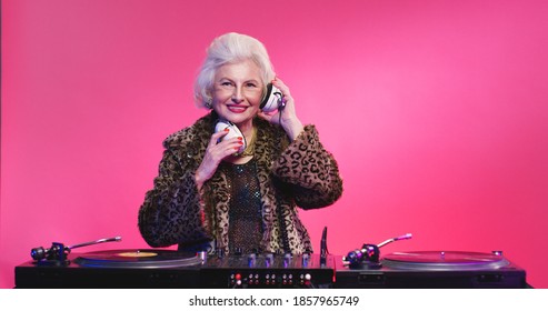 Cheerful and fancy vintage Caucasian grandmother in headphones and leopard coat while spinning music on mixer as DJ on retirement. Party of pensioners concept. Female having fun working at disco.