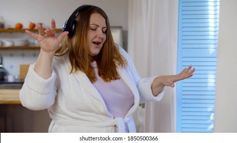 Cheerful Fat Woman Listening To Music In Headphones And Dancing Near Window At Home. Happy Overweight Lady In Bathrobe And Headset Enjoying Music In Morning