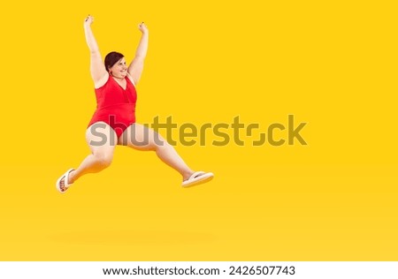 Cheerful fat woman in swimsuit rejoices, jumps and has fun while enjoying summer vacation. Funny chubby woman in red swimsuit hangs in air near copy space on yellow background. Banner. Full length.