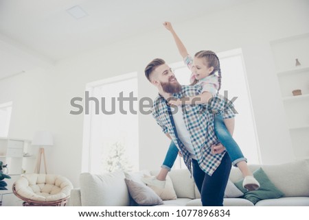 Cheerful father carrying on back little playful joyful kid with raised fist celebrating victory in game, family with one parent spending time in modern house with interior, babysitting concept
