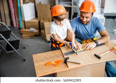 Cheerful father is helping little boy to use saw and hand tools on wooden desk in workshop