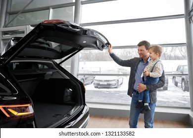 Cheerful Father Holding Kid On Hand, Showing To Son New Black Car. Handsome Man Opening Empty Car Trunk Of Expensive Black Automobile. Parent And Child Smiling, Looking At Car.